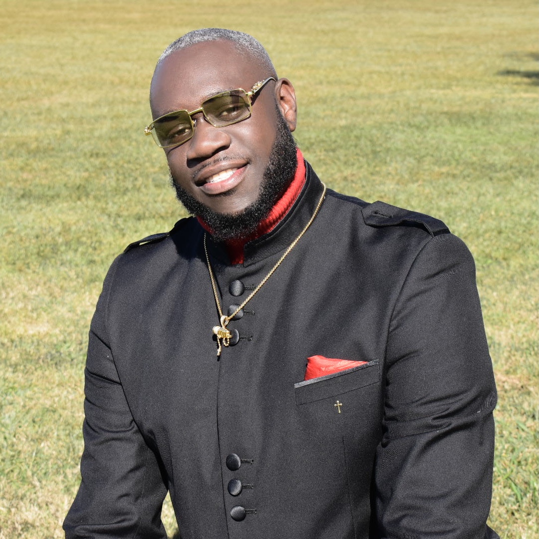 Pastor Albert L. Polk IV outdoors on a grass field in a black clergy suit with red accents